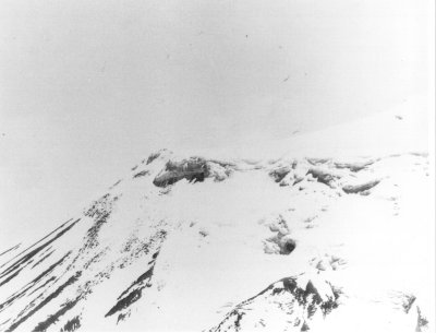 A grainy black and white aerial photograph of a snow-covered mountain ridge, with a darker roughly rectilinear patch in the ice near the crest.