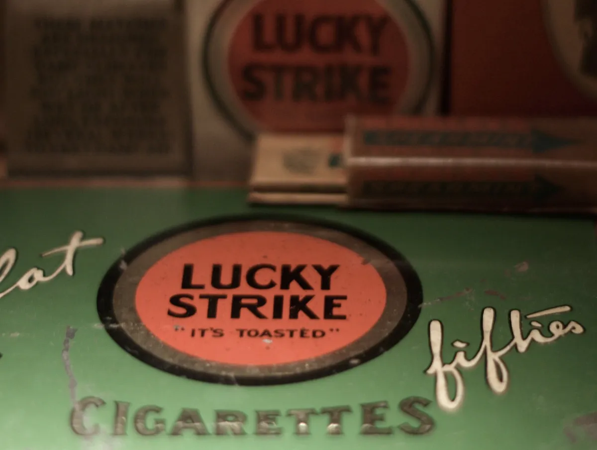 A green Lucky Strike cigarette tin marked "It’s Toasted", with white Lucky Strike packs behind it, displayed in a museum case.