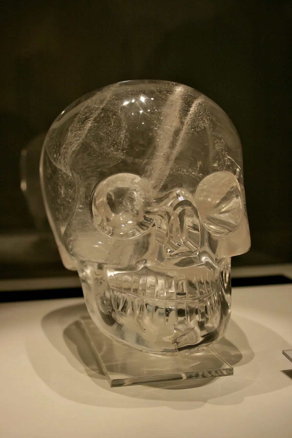 A life-size human skull carved from clear quartz, lit from below in a museum case, with rounded eye sockets and an evenly ruled row of teeth.