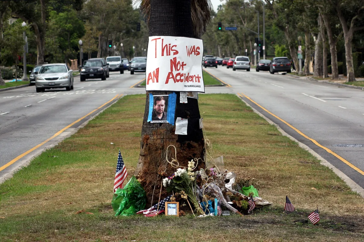 A palm tree on a road median in Los Angeles with flowers, small American flags and a photograph at its base, and a hand-lettered sign reading "This Was Not An Accident" fixed to the trunk.