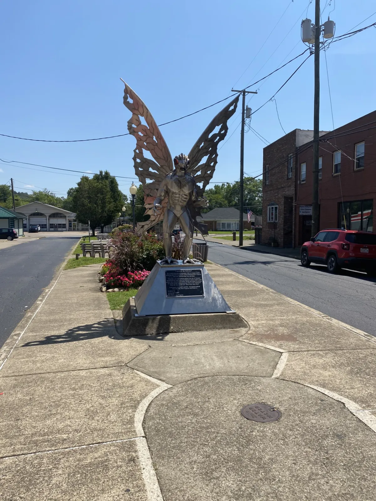A photograph of the Mothman Statue and surrounding street in Point Pleasant West Virginia. The statue is large and takes up most of the median, with streets on either side. The Mothman is a cryptid well known in the Midwest for his supposed involvement with the Silver Bridge collapse in Point Pleasa