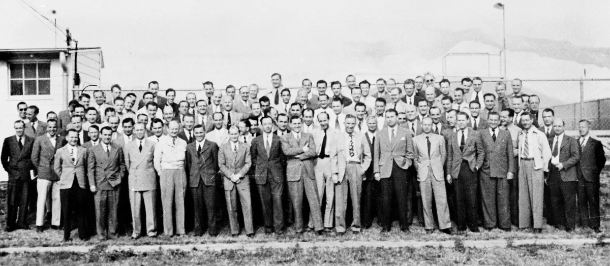 Rows of more than a hundred men in civilian suits posed outside a low building in open desert country.