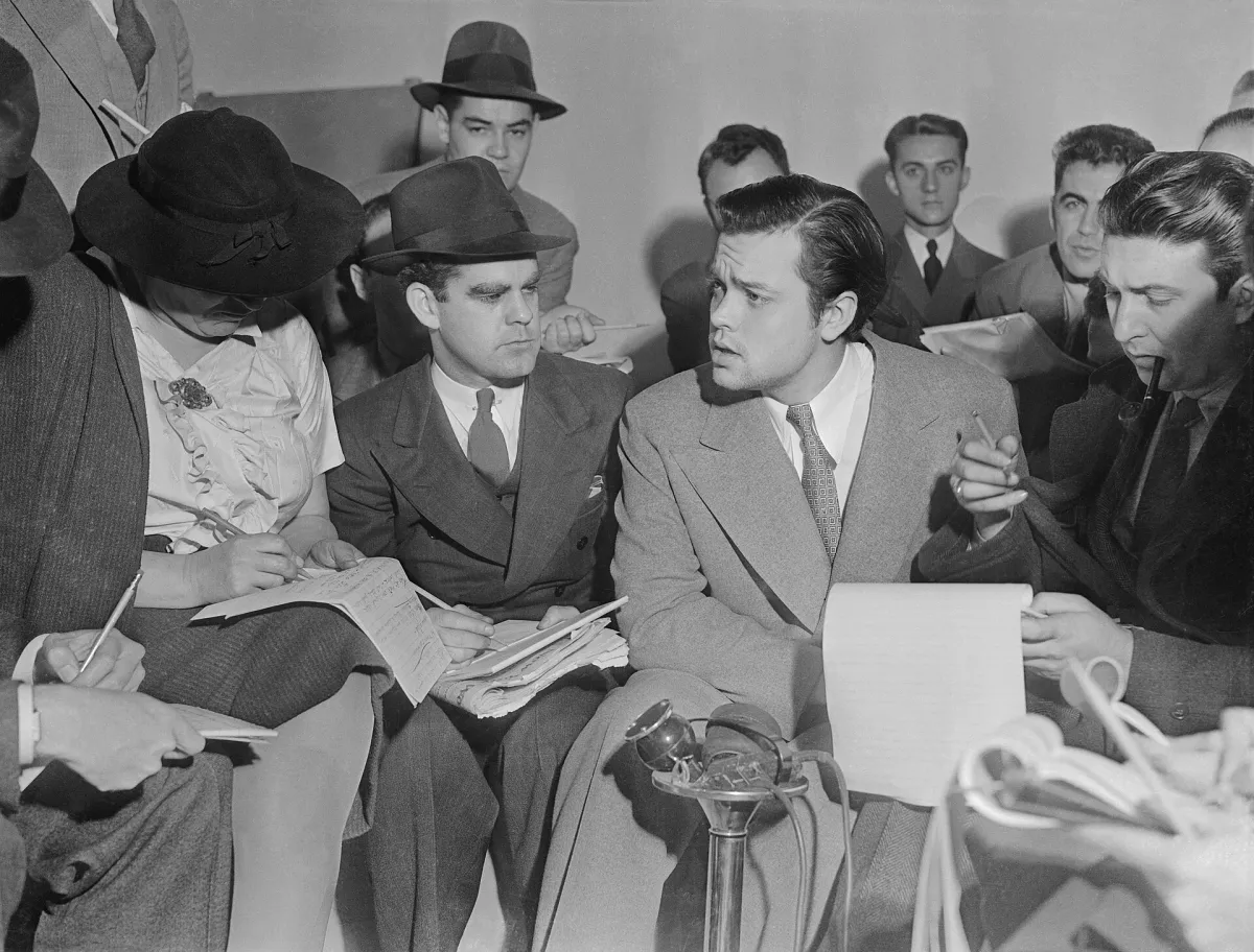 Orson Welles seated among a crowd of reporters who are taking notes, a microphone in front of him.