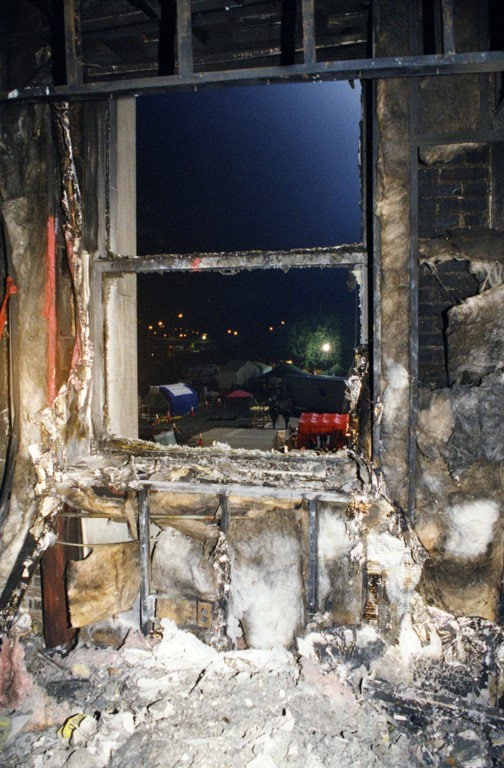 Night view through a burned-out window frame on an upper floor; response tents lit outside.