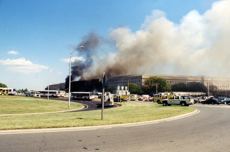 The west face seen from a nearby roadway, smoke still rising; buses and emergency vehicles in the foreground.