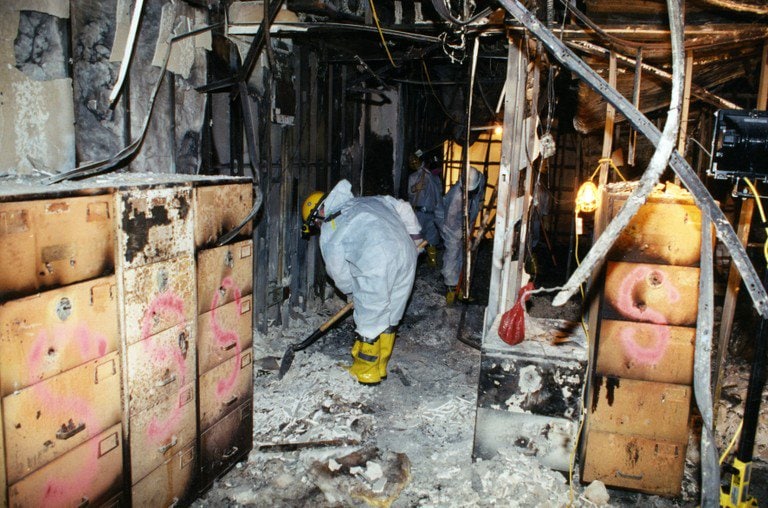 Interior office space, a worker in protective coveralls among burned filing cabinets marked in spray paint.