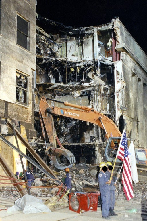 An excavator working at the collapsed facade after dark; workers with a flag in the foreground.