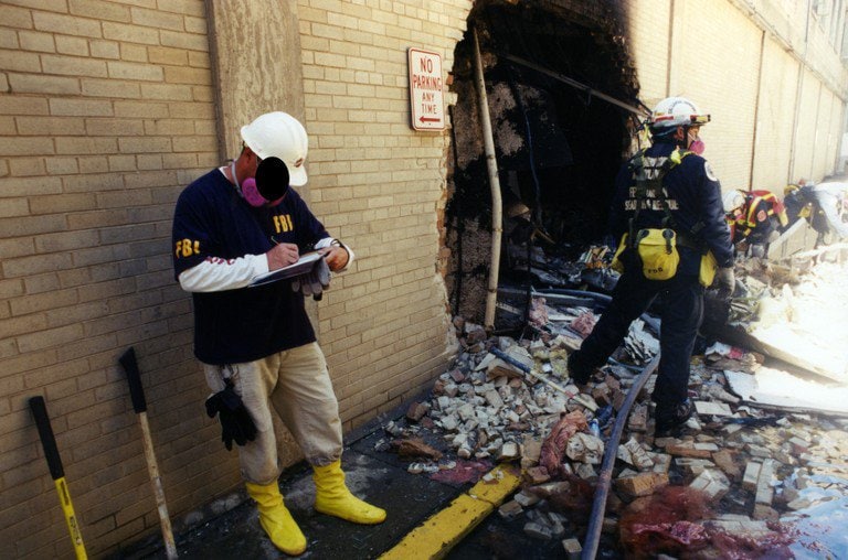 An investigator in an FBI jacket taking notes beside a breach in the exterior wall.