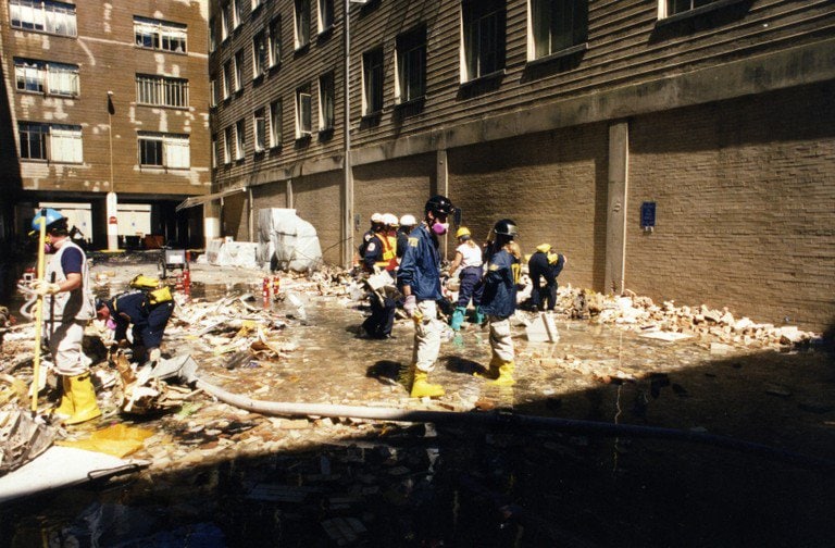 Workers on a debris-covered surface beside the building, standing water in the foreground.