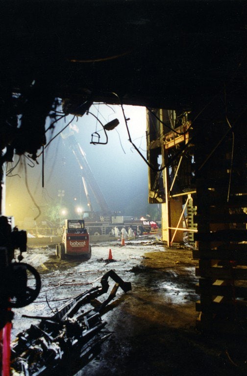 Looking out through a breach in the outer wall at night, a crane and a skid-steer loader working under floodlights beyond.