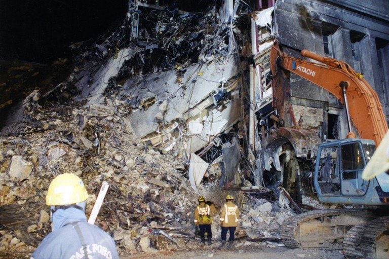 The collapsed section under floodlights, an excavator at the right and firefighters at the foot of the debris slope.