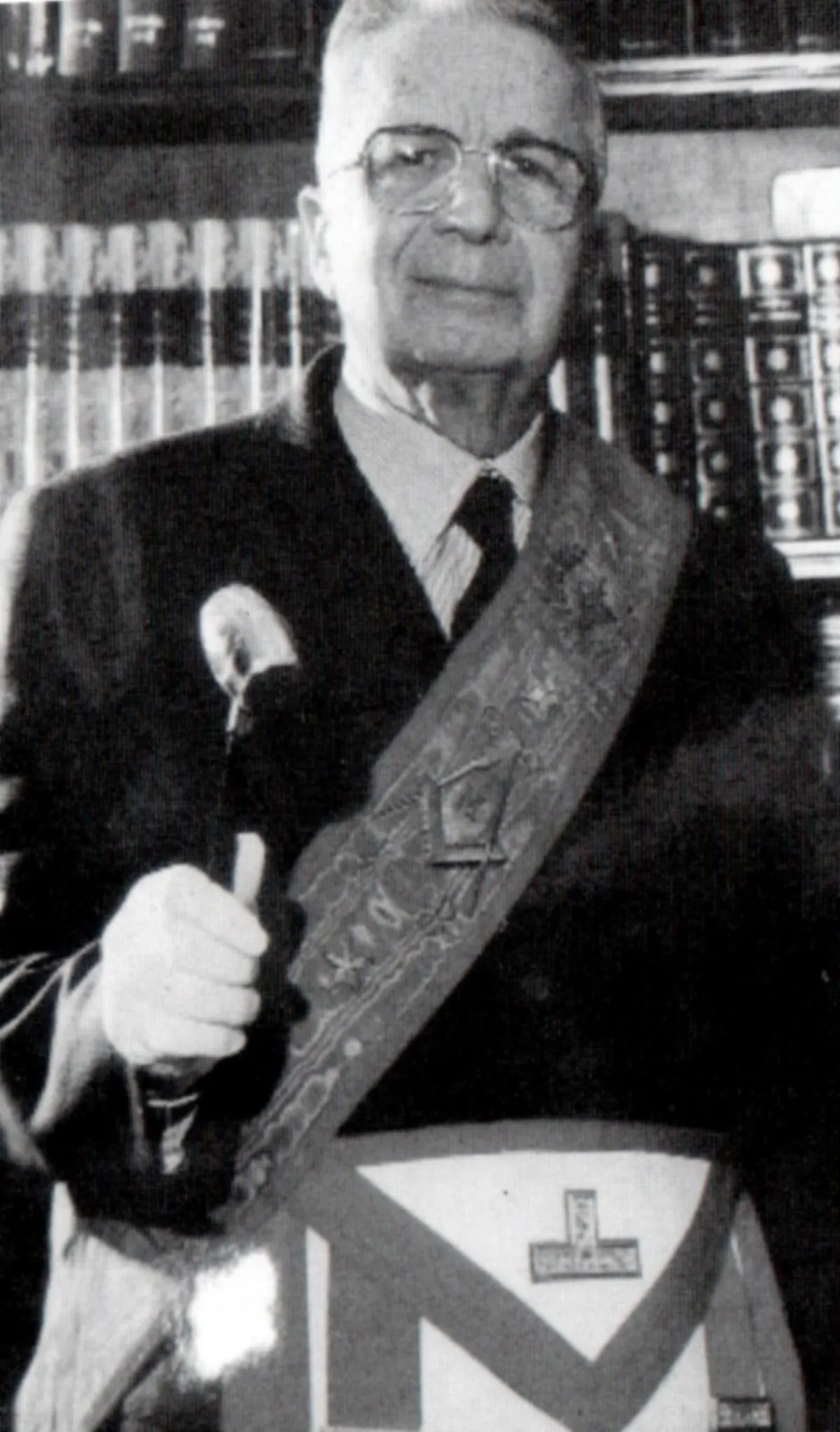 Licio Gelli, an elderly man in glasses, wearing a masonic sash and apron and holding a gavel, in front of shelves of books.
